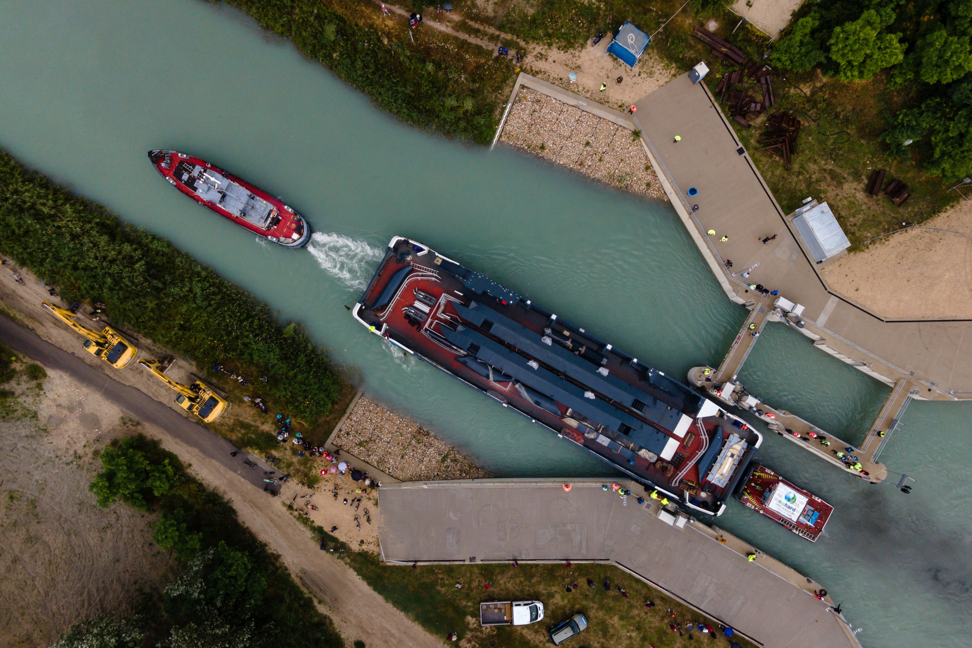 A drónnal készült felvételen a Balatoni Hajózási Zrt. (Bahart) egyik új kompját vontatják a Sió-csatorna balatonkiliti duzzasztógátjánál 2023. június 28-án. A Bahart a Magyar Turisztikai Ügynökség (MTÜ) közreműködésével építtetett két új személyhajója és két kompja 2021-ben készült el a komáromi hajógyárban, ahonnan egy héttel ezelőtt indultak útnak, hogy a Dunán lehajózva a Sió-csatornán keresztül elérjék a Balatont. 